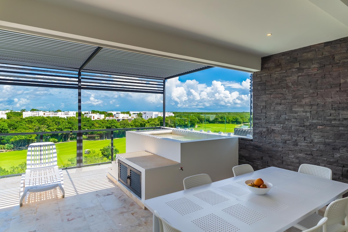 A spacious outdoor dining area is showcased with a white table and colorful fruit centerpiece. A built-in grill is present alongside a splash pool, with panoramic views of lush greenery and distant white buildings visible through expansive glass sliding doors.