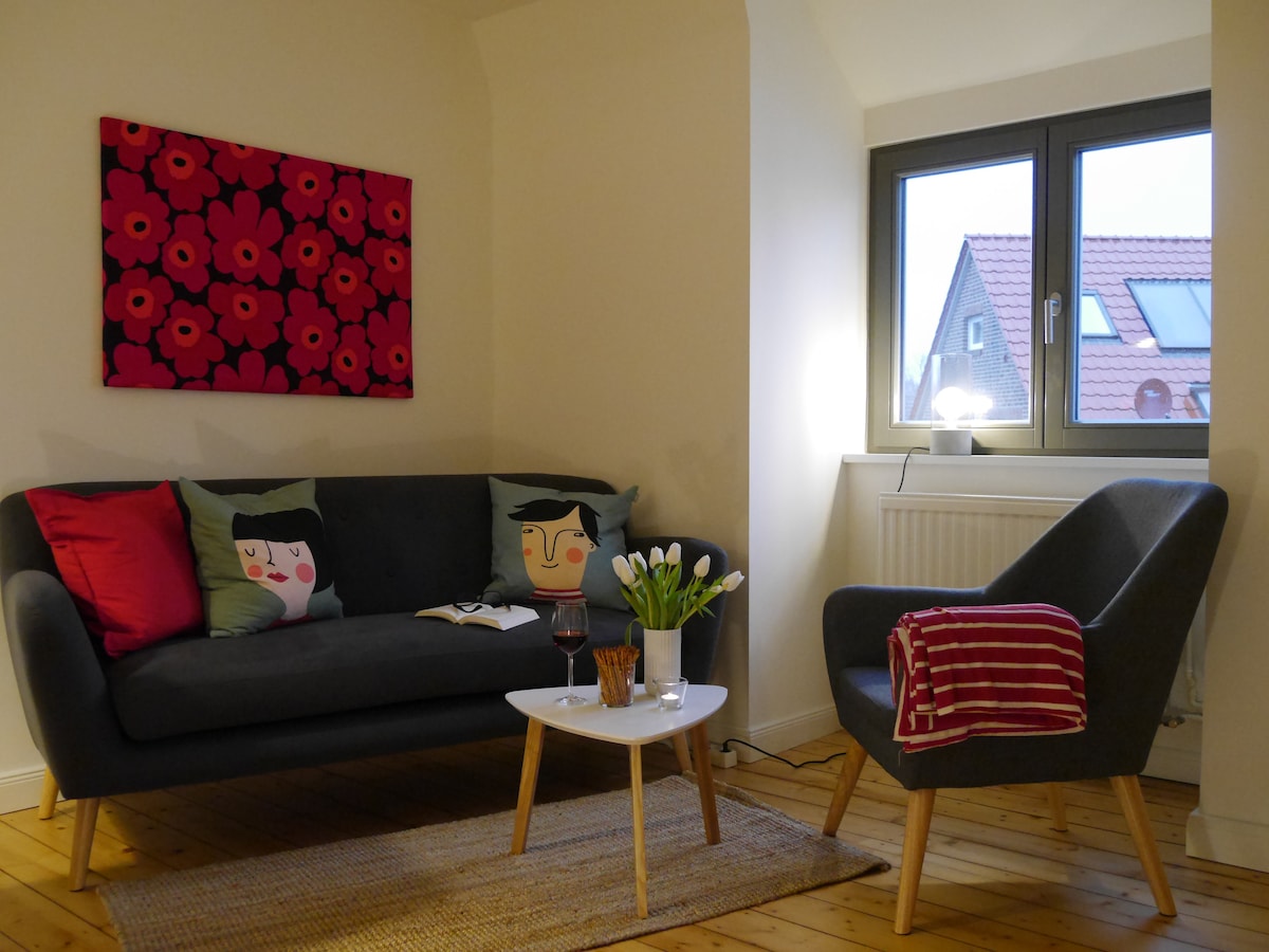A cozy living area features a grey sofa adorned with decorative cushions, alongside a stylish armchair. A small table is set with a glass of wine and a bouquet of white tulips. Natural light is filtered through the window, enhancing the welcoming ambiance.