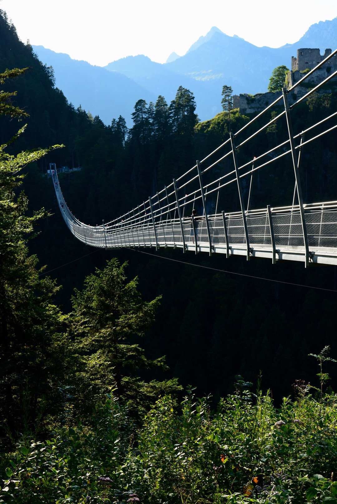 A suspension bridge stretches gracefully across a valley, surrounded by lush greenery and towering trees. In the background, rugged mountains are visible, with the silhouette of a castle perched on a hillside, enhancing the dramatic landscape.