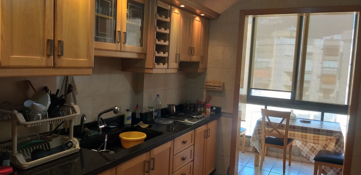 A kitchen featuring wooden cabinetry and countertop space is shown. Cooking appliances and utensils are neatly arranged, with a visible sink and drying rack. A dining table with chairs is positioned beside a large window, allowing natural light to illuminate the space.