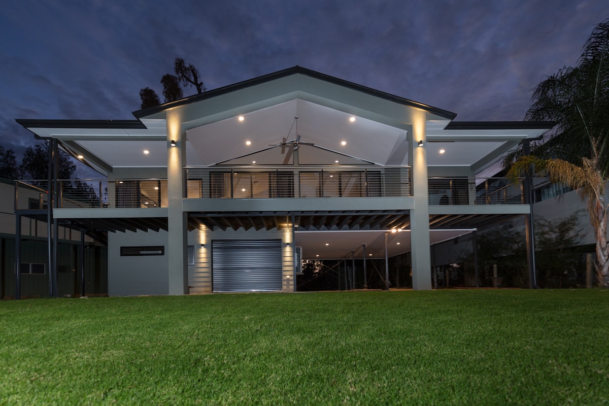 The modern riverfront home is shown at twilight, illuminated by exterior lights. A spacious lawn leads up to the elevated structure with a prominent gabled roof and expansive deck. Large glass doors reveal the interior, blending contemporary design with the natural surroundings.
