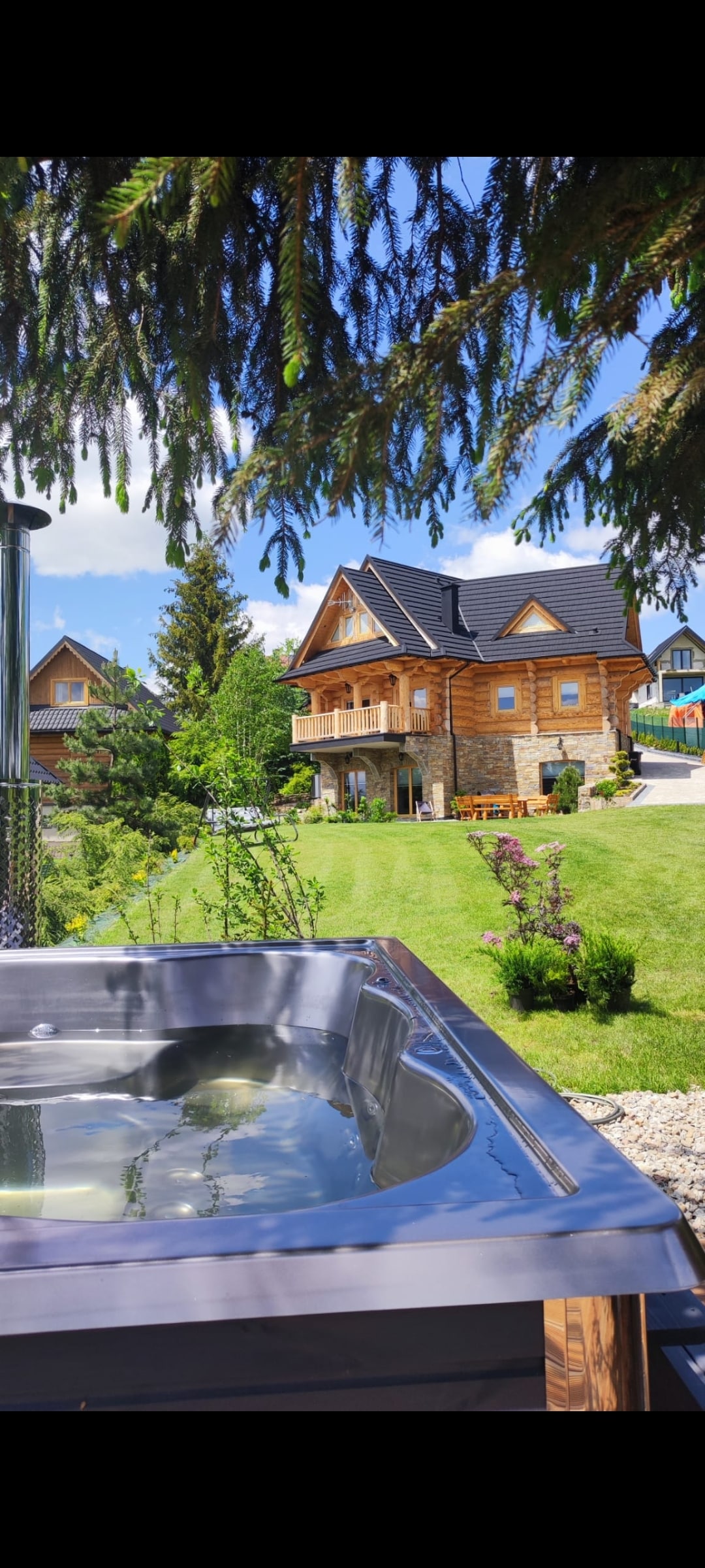 A spacious wooden house with an elegant design is set against a backdrop of green trees and mountains. A hot tub is prominently featured in the foreground, with clear water inviting relaxation. A well-maintained garden surrounds the property, enhancing its natural appeal.