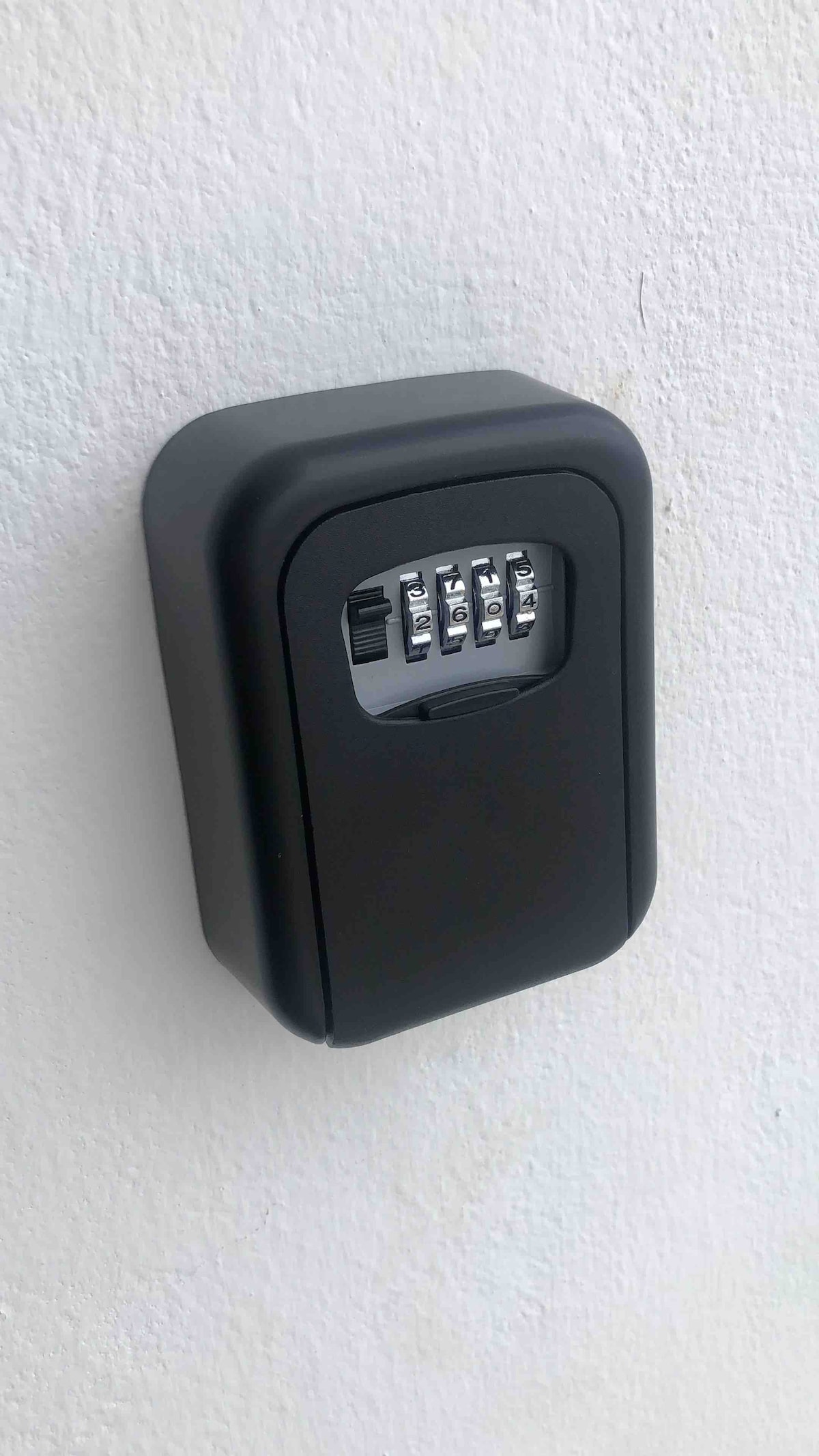 A secure key lockbox is mounted on a white wall, featuring a combination dial with five numeric buttons. The lockbox is made of black material, designed to provide easy and secure access to keys.