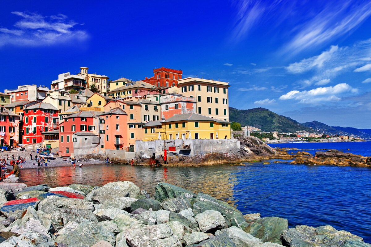 A colorful array of buildings lines the coastline, reflecting classic architectural styles. The scene includes rocky formations in the foreground, leading to a calm blue sea. The backdrop features gentle hills under a bright sky, complemented by scattered clouds.
