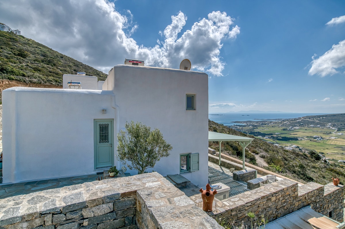 A newly built cycladic home is visible, featuring clean white walls and a green door. A stone pathway leads to a covered outdoor seating area with views of the landscape, while hills and the sea can be seen in the background, complemented by a partly cloudy sky.