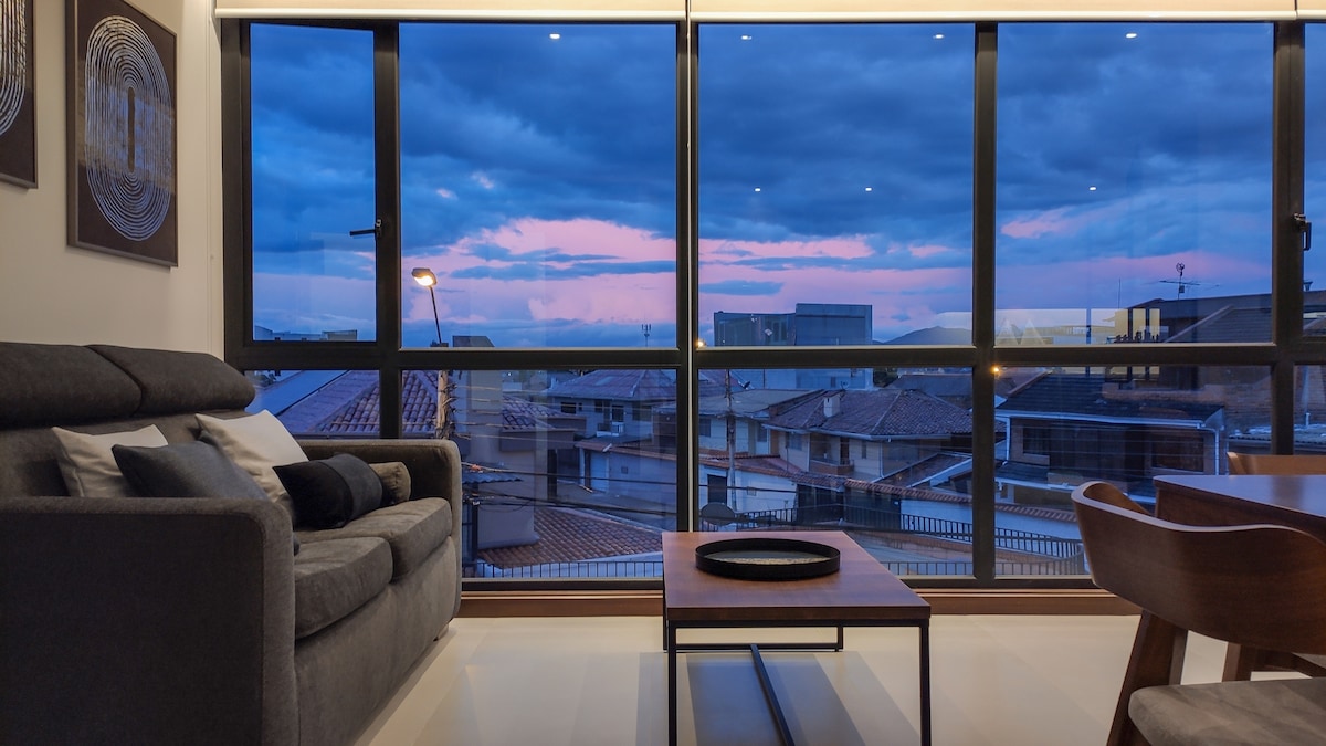 The living area features large floor-to-ceiling windows offering a view of the city and sky during twilight. A comfortable sofa and a coffee table create a relaxing atmosphere, while the dining area with wooden chairs is softly illuminated by ambient light.