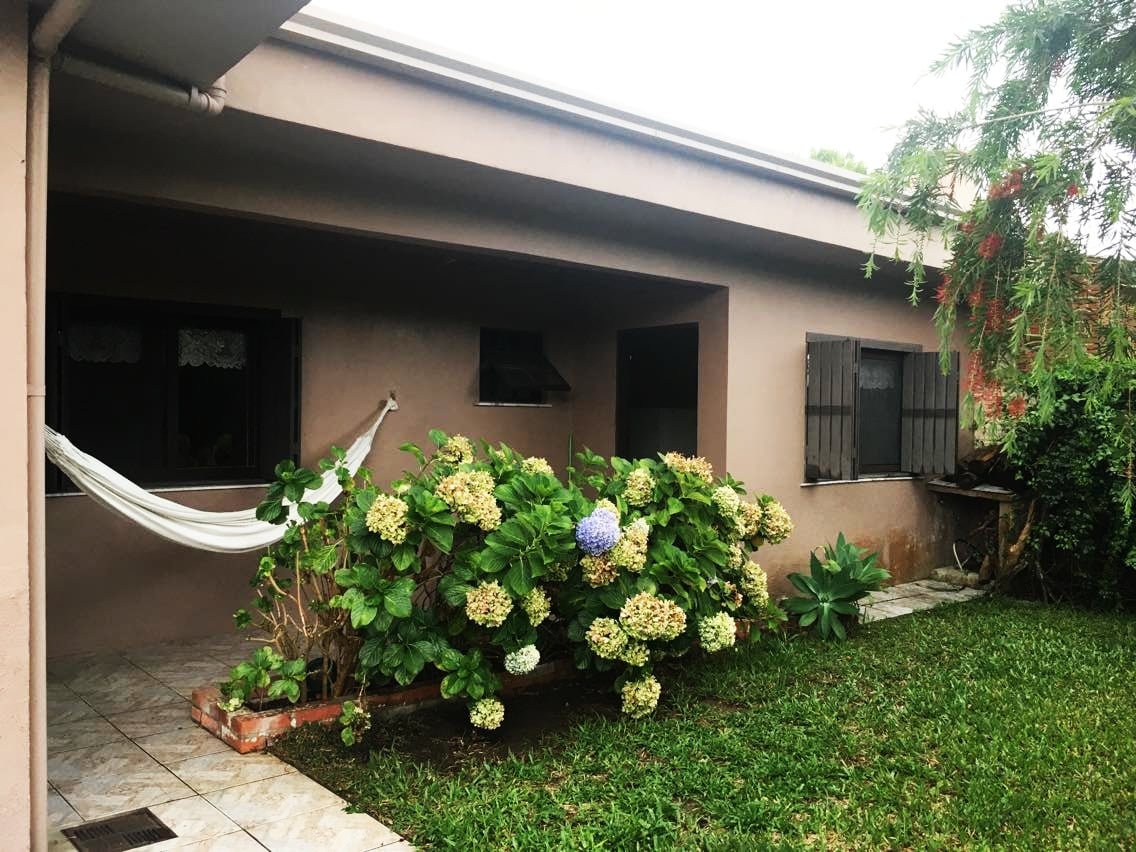 A welcoming exterior view of the house features a shaded patio area surrounded by lush greenery and flowering plants. A hammock is seen suspended between two points, inviting relaxation. Natural light fills the space, with windows allowing a glimpse into the indoors.