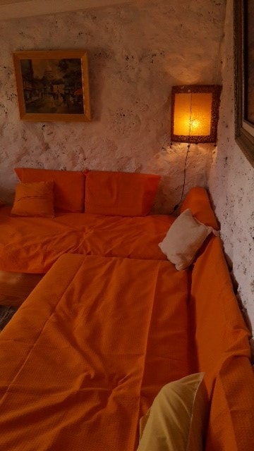 A cozy seating area features a spacious orange couch adorned with several cushions of varying sizes. A soft, warm light emanates from a bedside lamp, illuminating the textured walls and enhancing the comfortable ambiance.