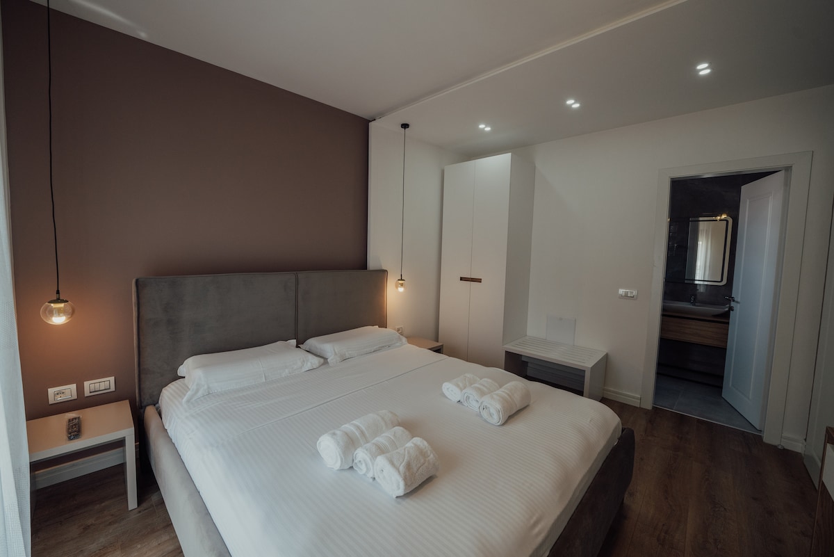 A modern King room features a spacious bed with neatly arranged towels, complemented by soft lighting from pendant fixtures. A wardrobe and a bedside table are positioned beside the bed. A private bathroom doorway is visible in the background, enhancing the room's functionality.
