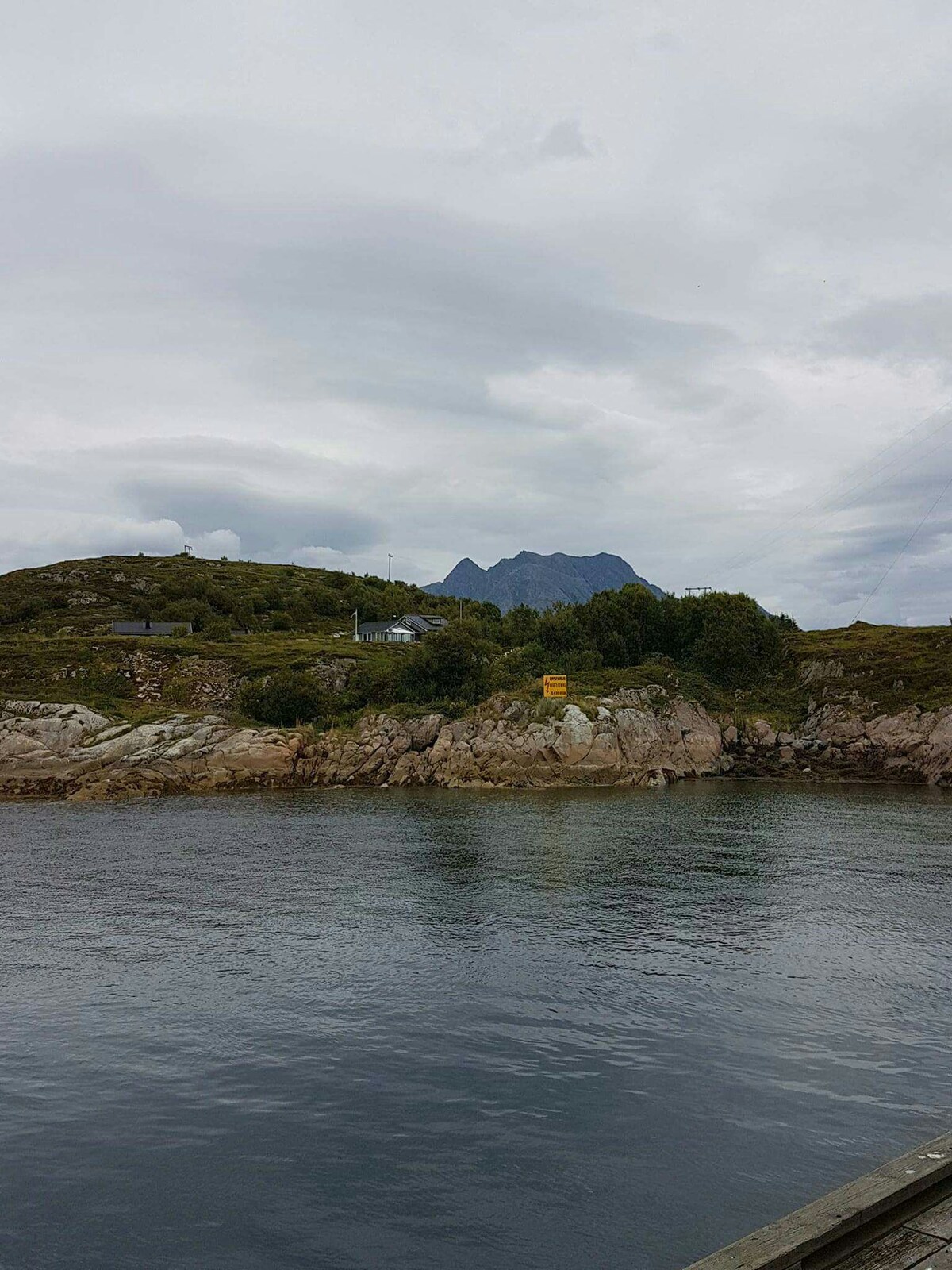 A serene view of a rocky shoreline meets calm waters, with distant hills rising in the background. Overcast skies cast a soft light over the landscape, highlighting the natural beauty of the surroundings.
