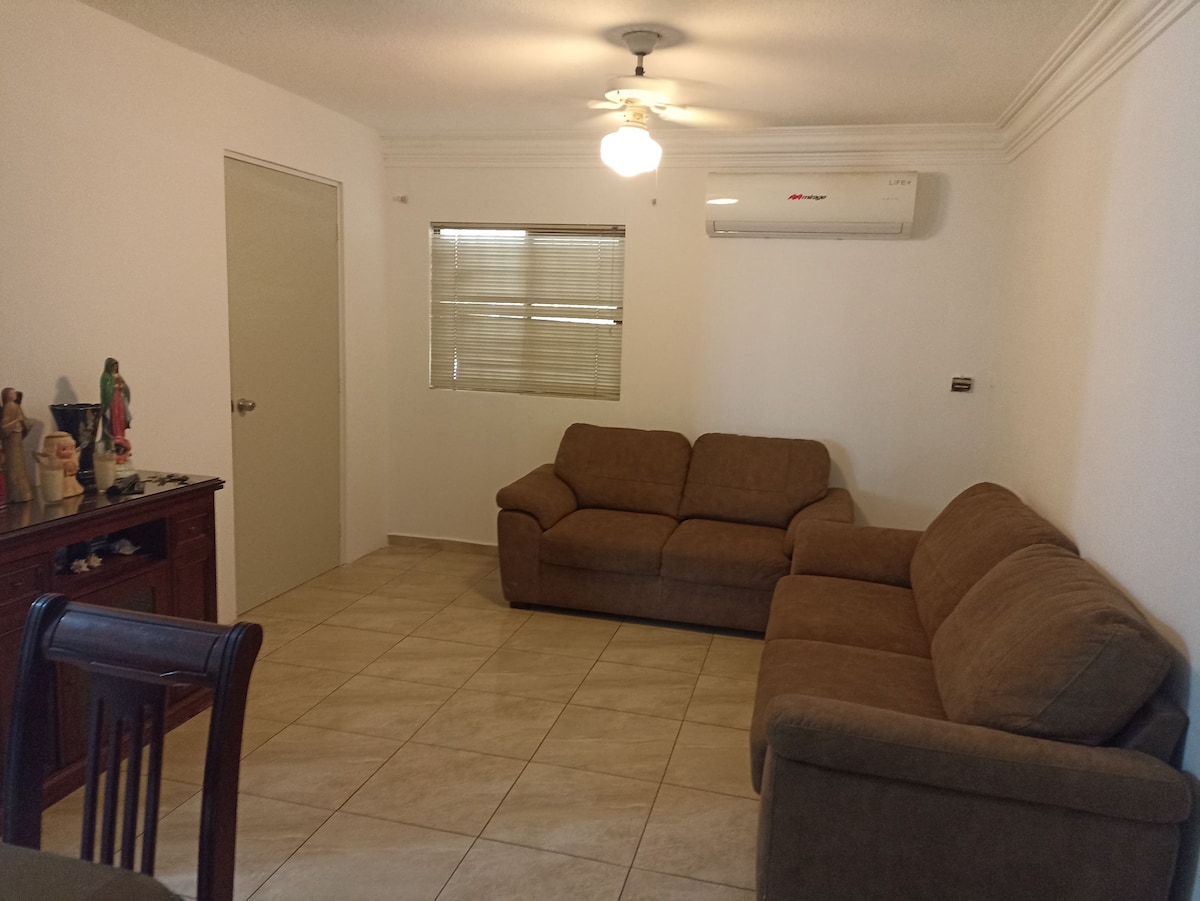 A comfortable living area features two tan sofas, a ceiling fan, and an air conditioning unit. Light-colored tiles floor the space, while a wooden console table displays decorative items against a wall. Natural light enters through a window with blinds.