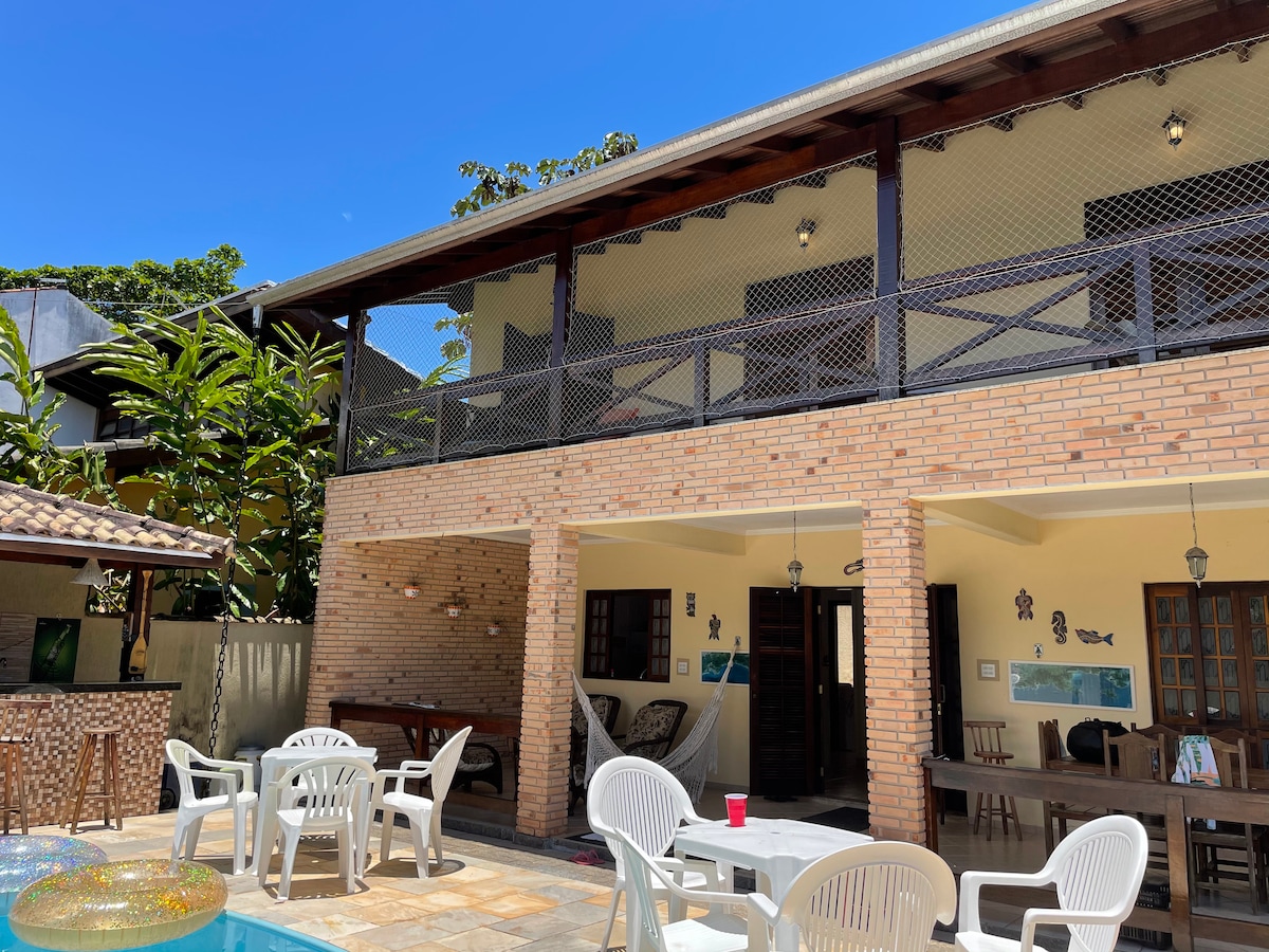 The two-story house features a brick facade with an open terrace above, complemented by wooden beams. A spacious outdoor area includes a table and white chairs. Lush greenery surrounds the property, and bright blue skies provide a vibrant backdrop.