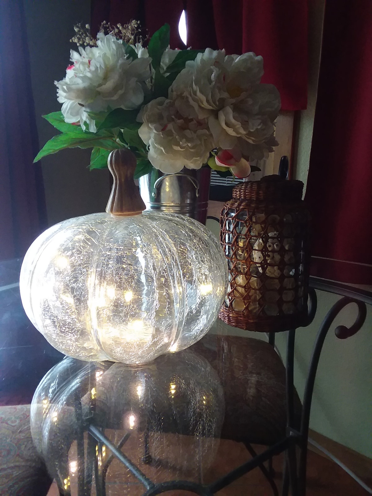 A decorative glass pumpkin, illuminated from within, rests on a reflective table. Fresh flowers in a vase and a textured lantern add to the cozy ambiance of the space, complemented by the rich, red curtains in the background.