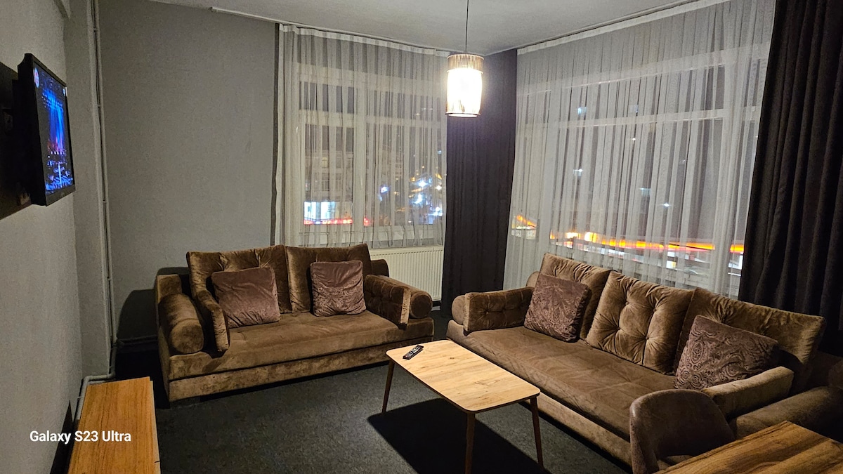 A spacious living area features two comfortable sofas arranged facing a wooden coffee table. Large windows with sheer curtains provide a view of the street, while a flat-screen TV is mounted on the wall, enhancing the room's functionality.