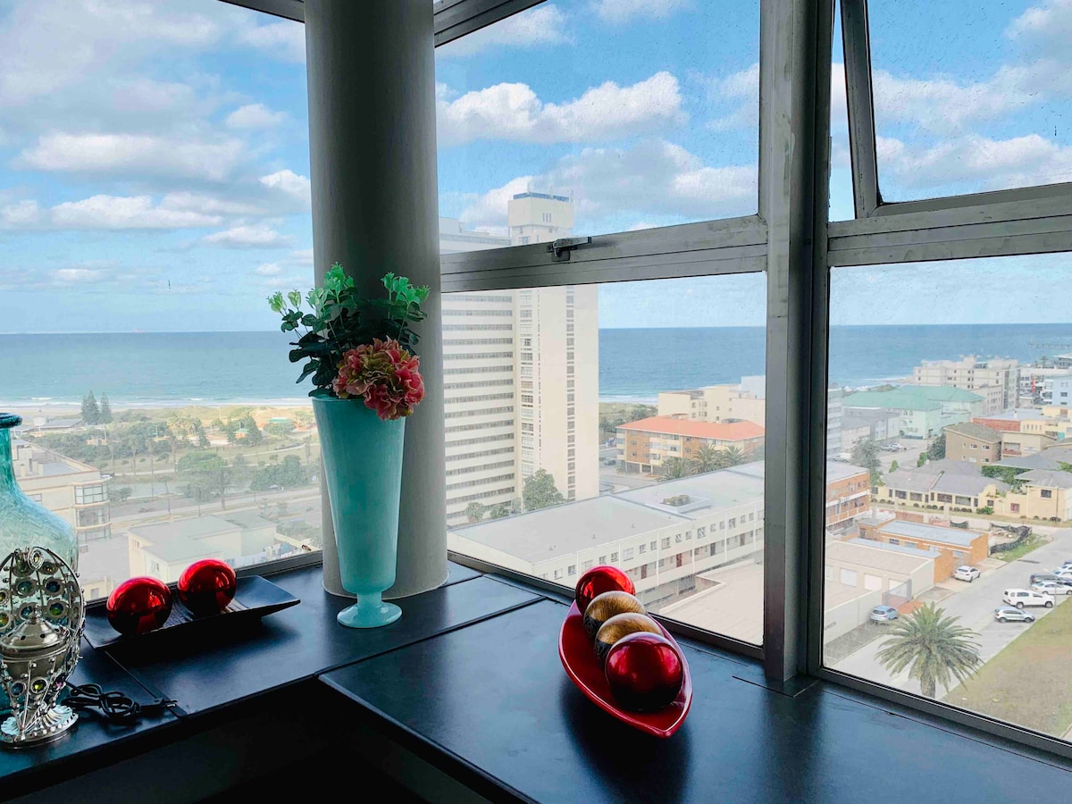 A large window offers a panoramic view of the sea and surrounding landscape. Decorative elements include a blue vase with flowers and a decorative bowl in red, adding a touch of color to the dark surface below.