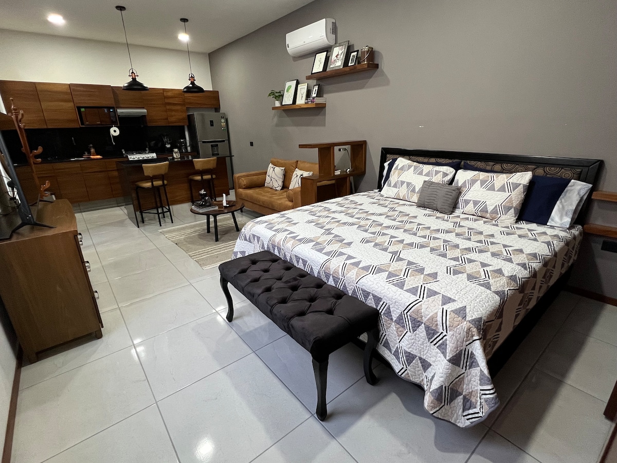 A spacious loft features a large bed with a patterned bedspread, complemented by a tufted bench at its foot. A cozy seating area with a light brown sofa is visible, along with a small dining set and modern kitchen cabinetry in the background.