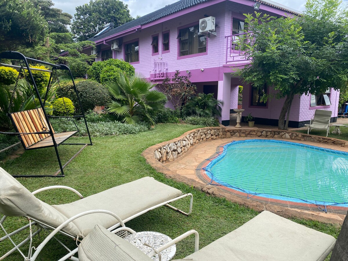 A lush garden area features a small pool with a turquoise surface, surrounded by stone edging. Two lounge chairs are positioned nearby, alongside a wooden swing set. The vibrant exterior of a two-story home is visible, enhanced by greenery and shaded by trees.