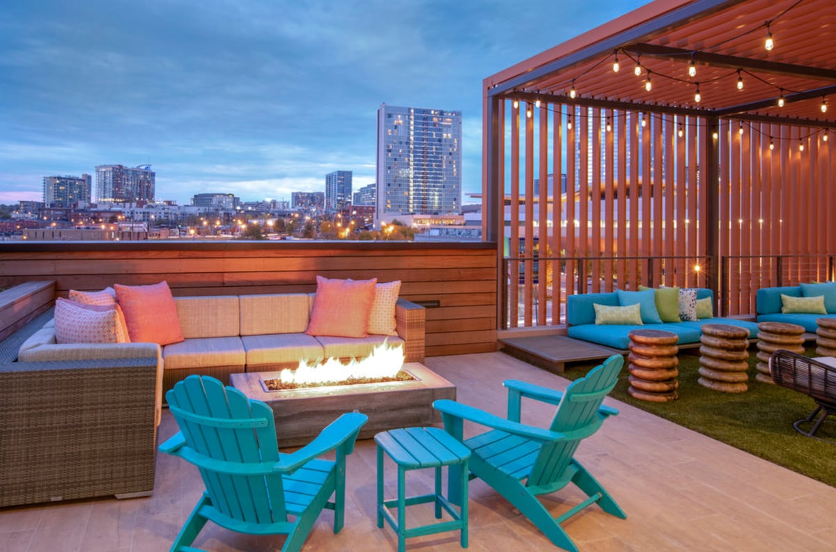 A rooftop terrace features a modern seating area that includes a sectional sofa with colorful pillows and turquoise chairs. A fire pit is centrally located, surrounded by stacked wooden stools. Strings of lights above provide a soft glow, while a city skyline is visible in the background.