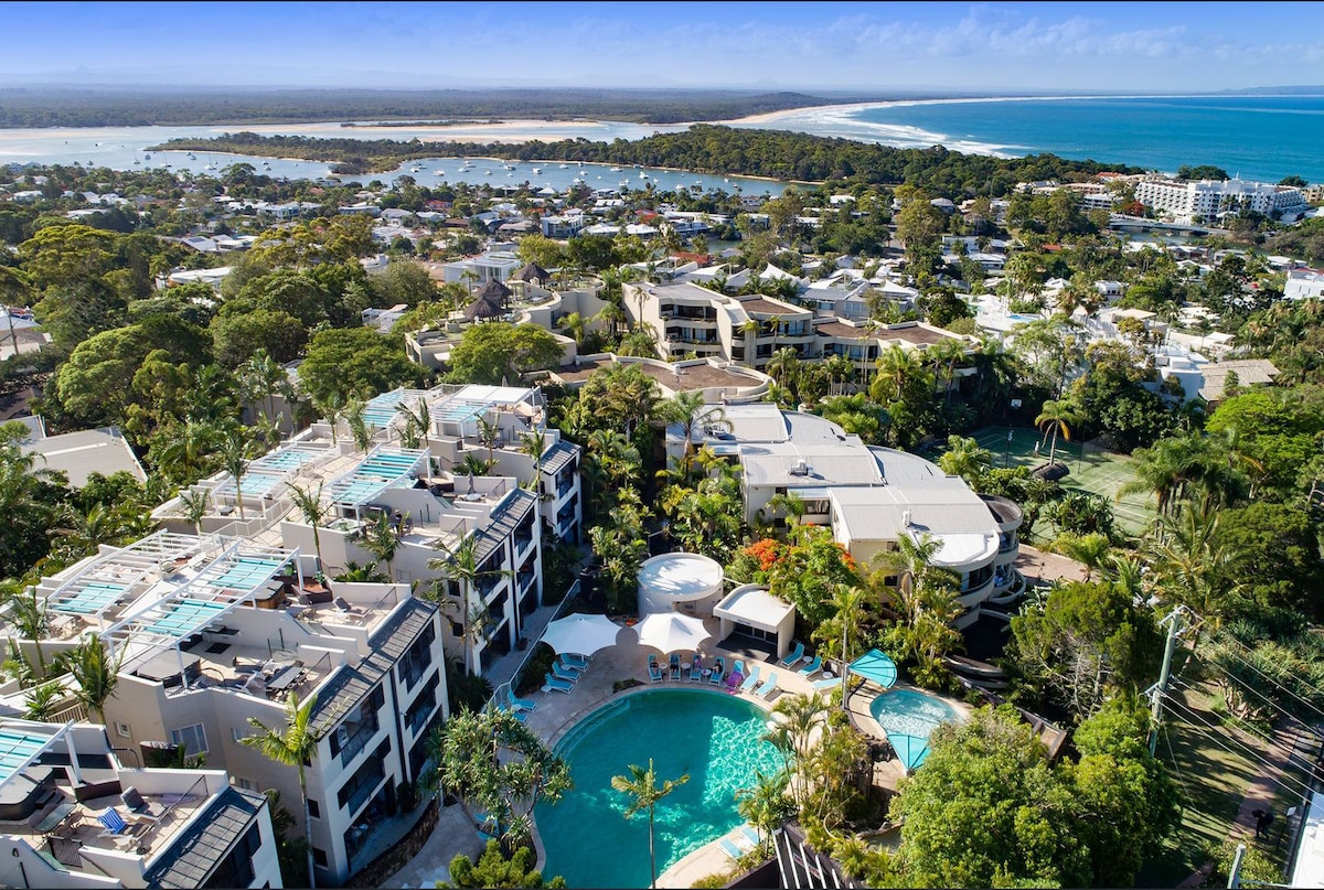 Aerial view of a resort complex surrounded by lush greenery and palm trees, featuring a central pool area with lounge chairs. The coastline and water are visible in the distance, illustrating a serene environment close to nature. Buildings showcase modern architecture.