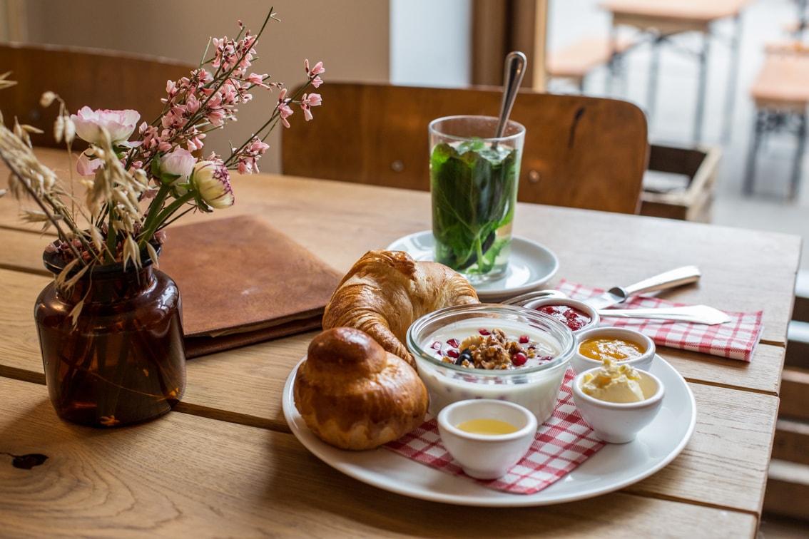 A wooden table is set with a plate of breakfast items including a croissant, a sweet pastry, yogurt topped with granola and berries, along with small bowls of honey and butter. A vase with flowers and a drink sits nearby, creating a charming dining atmosphere.