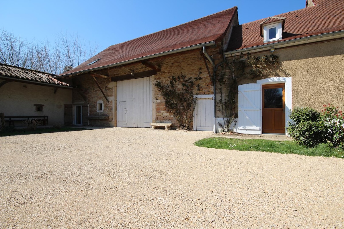 A spacious courtyard area is shown, featuring a gravel surface and flanked by a rustic stone building with a large wooden door. Sunlight illuminates the setting, and greenery is visible along the edges, creating a peaceful outdoor environment.