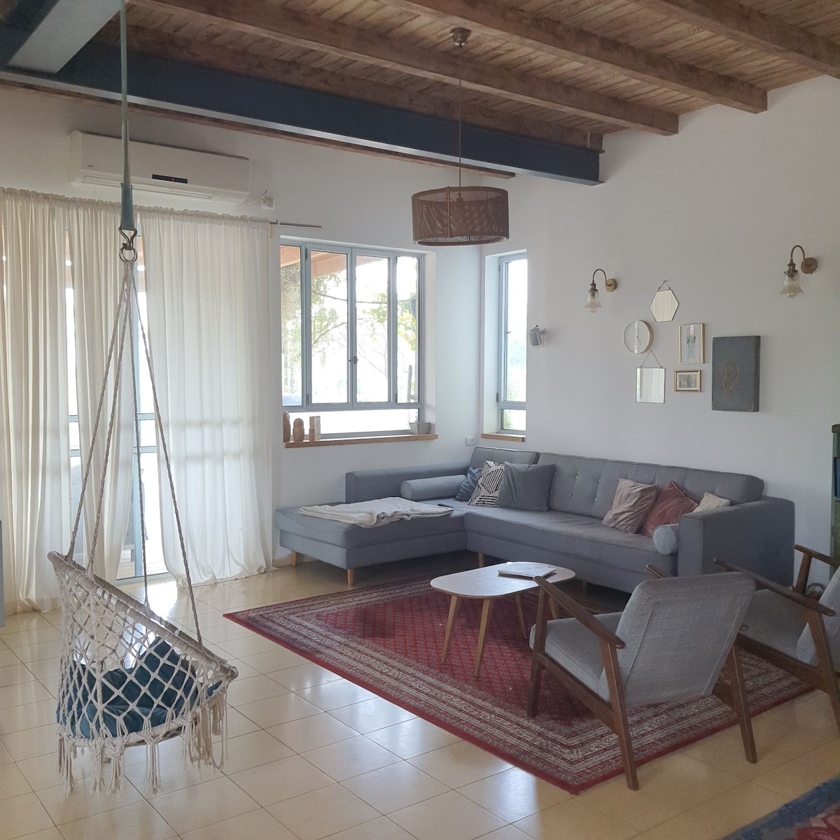 A spacious living area features a large sectional sofa and armchairs, positioned on a colorful area rug. Natural light streams through large windows, enhancing the open feel. A hanging chair adds an element of relaxation, while decorative wall mirrors and art pieces provide subtle visual interest.