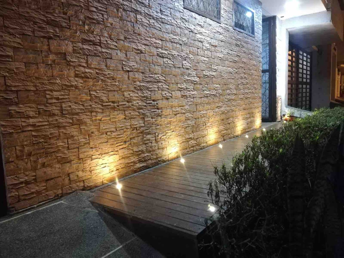 An exterior view of a stone wall illuminated by soft lighting fixtures along a wooden pathway. Lush greenery is visible on one side, enhancing the inviting ambiance of the entrance. The overall layout suggests a well-maintained entrance to the property.