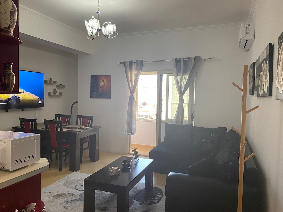 A modern living space is displayed, featuring a dark sofa accompanied by a decorative throw. A coffee table sits centrally on a soft rug, while a dining table with red-accented chairs is positioned nearby. A large window invites natural light, enhancing the room's ambiance.