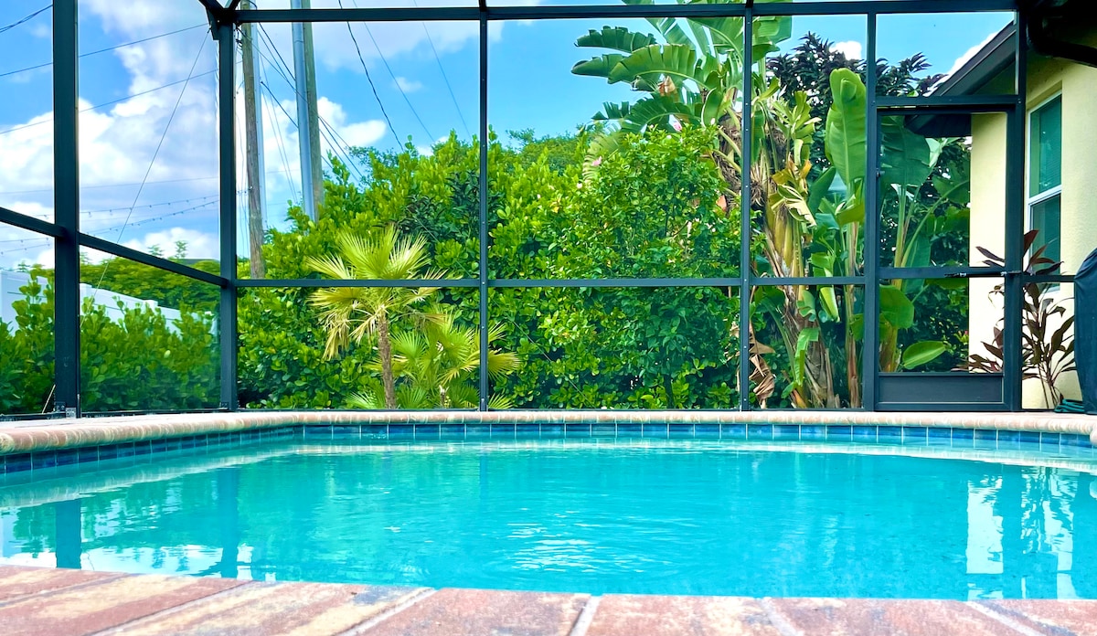 A tranquil swimming pool is framed by a screened enclosure, surrounded by lush greenery. Clear water reflects the blue sky, and the pool area features brick paving, providing a welcoming setting for relaxation.