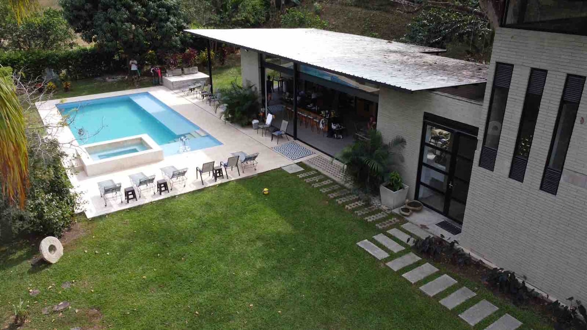 The outdoor space features a saltwater pool with an adjacent jacuzzi, surrounded by lounge chairs. A spacious open area is visible, connected to the indoor spaces through large glass doors. Lush greenery frames the scene, creating a serene atmosphere.