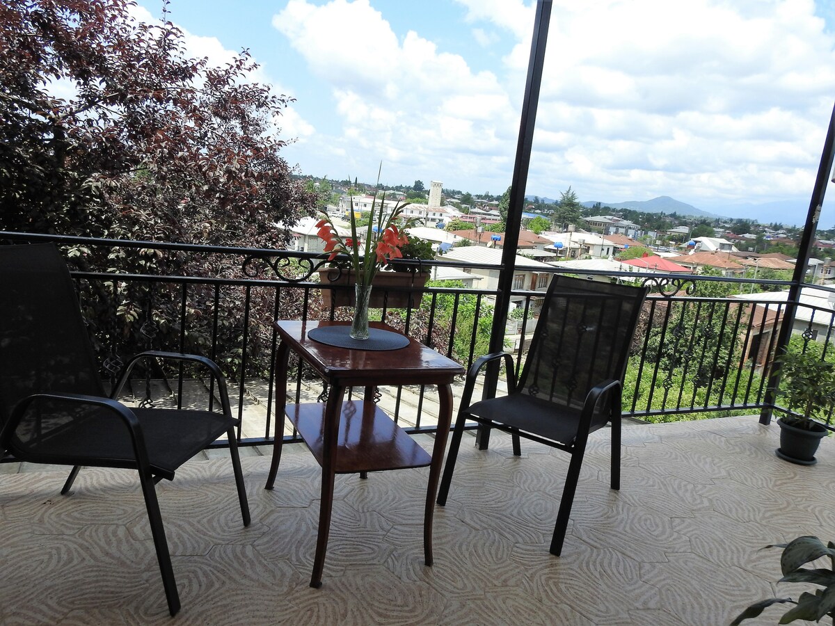 A covered terrace is presented with two black chairs and a small wooden table. A floral arrangement sits atop the table, while the surrounding greenery and distant rooftops are framed by a scenic view under a partly cloudy sky.