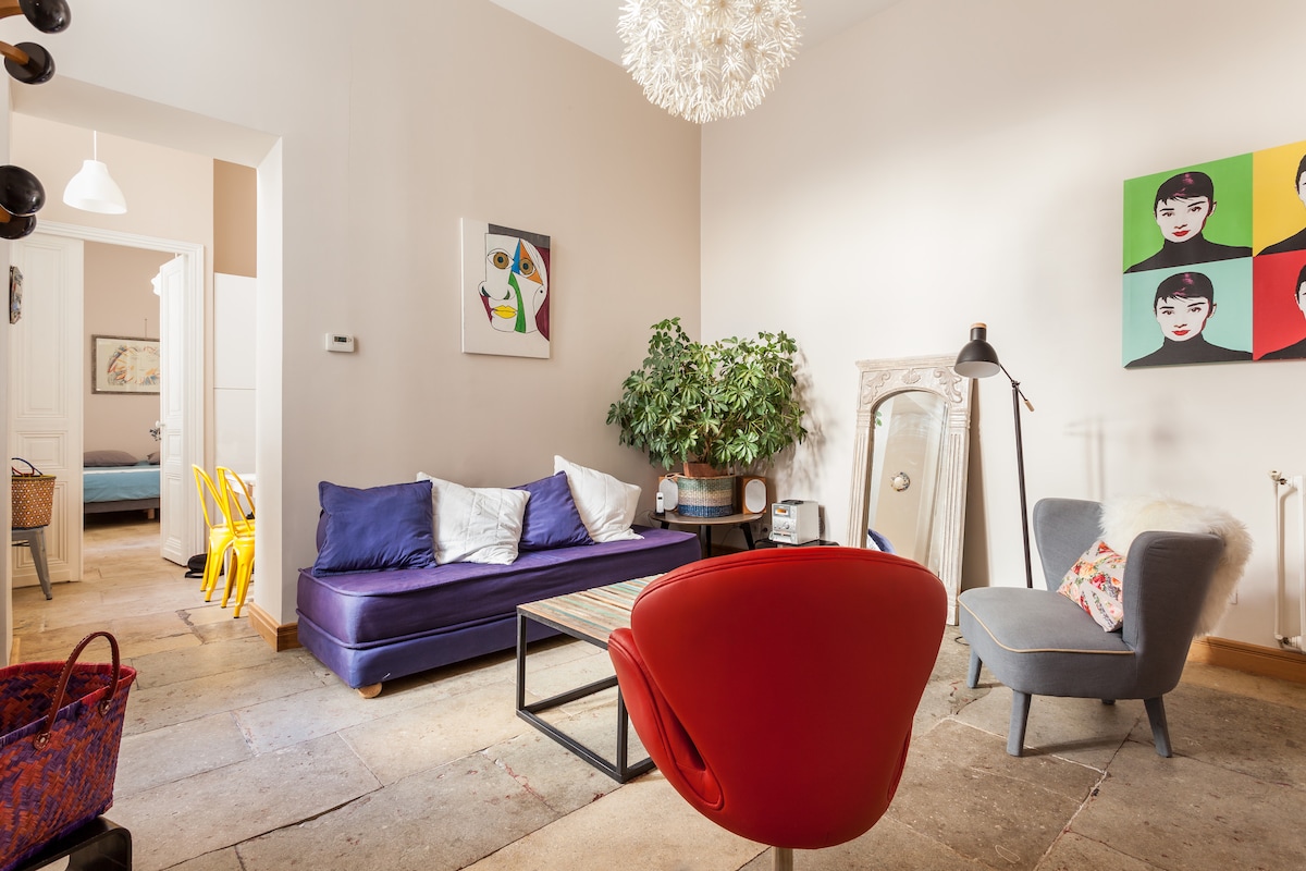The living area features a deep blue sofa adorned with decorative cushions. A colorful armchair adds a pop of red. A natural green plant enhances the space, while a wooden coffee table sits centrally, surrounded by light-colored stone flooring. Art pieces in vibrant hues adorn the wall.