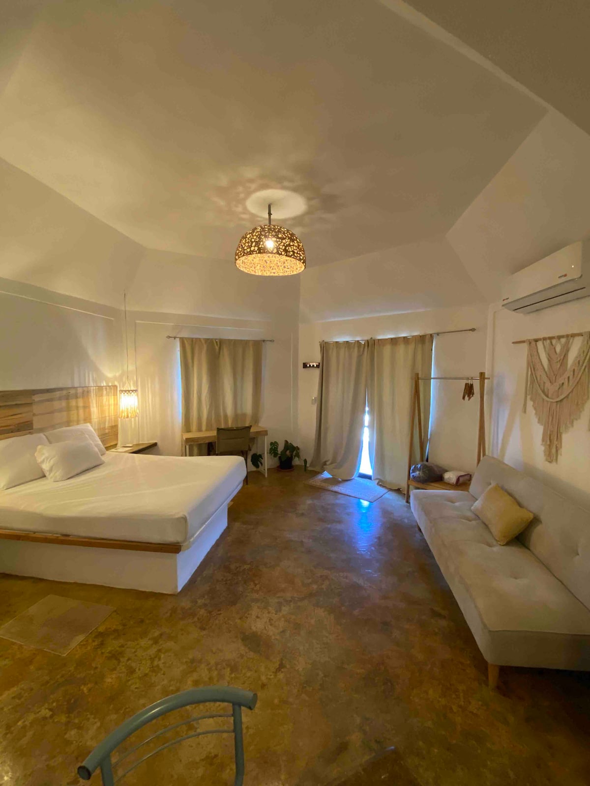 A spacious room features a large bed and a comfortable seating area. Natural light filters through multiple curtains, casting a warm glow on the stone flooring. A decorative light fixture hangs from the ceiling, while plants add a touch of green.