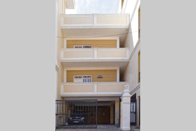 The exterior of the building is showcased, displaying a three-level structure with balconies and a gated entrance. A parking area is visible at the base, with a vehicle parked inside. Light colors and simple architecture characterize the façade, creating a welcoming appearance.