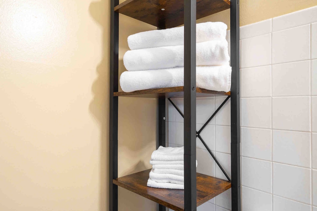 A sturdy wooden shelf displays neatly stacked white towels, emphasizing a clean and organized bathroom space. The backdrop features warm beige walls and a tiled section, creating a balanced and functional design.