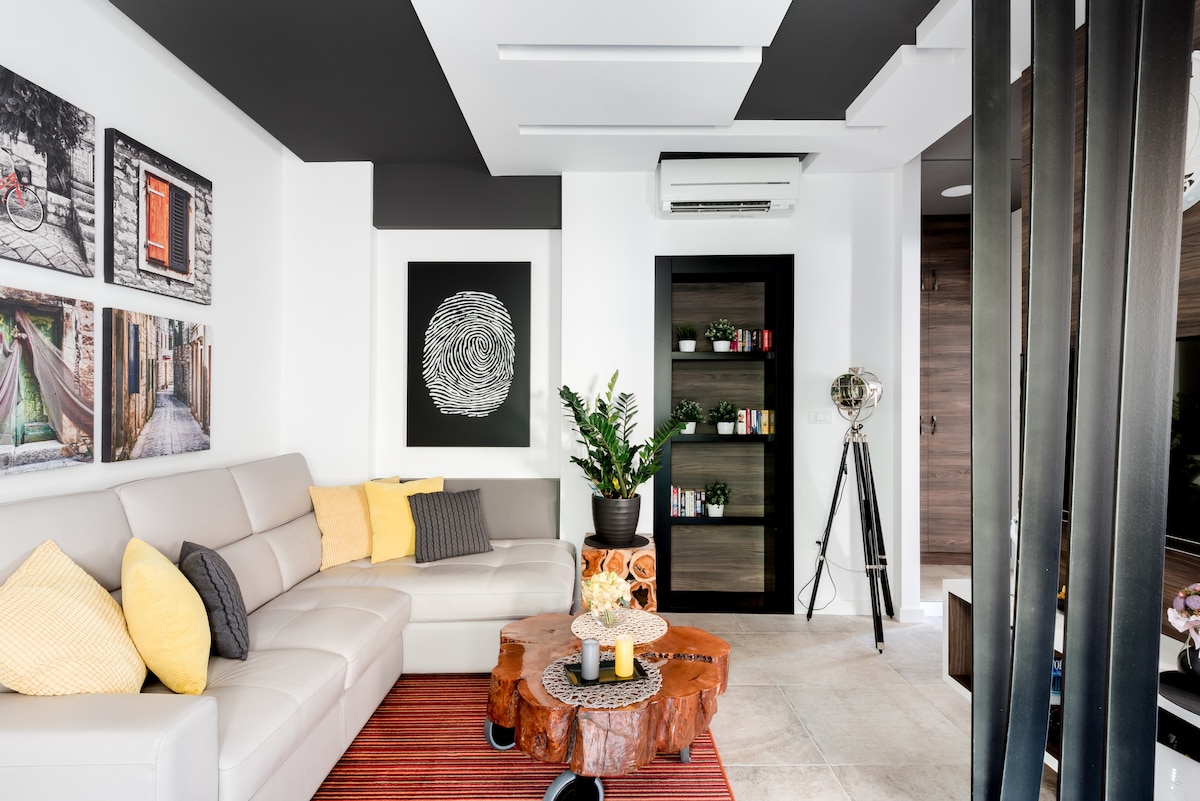 A modern living area features a light-colored sectional sofa adorned with yellow and gray pillows. A wooden coffee table with decorative items is centered on the textured rug. Artwork adorns one wall, and a sleek bookshelf holds various objects, enhancing the space's contemporary design.