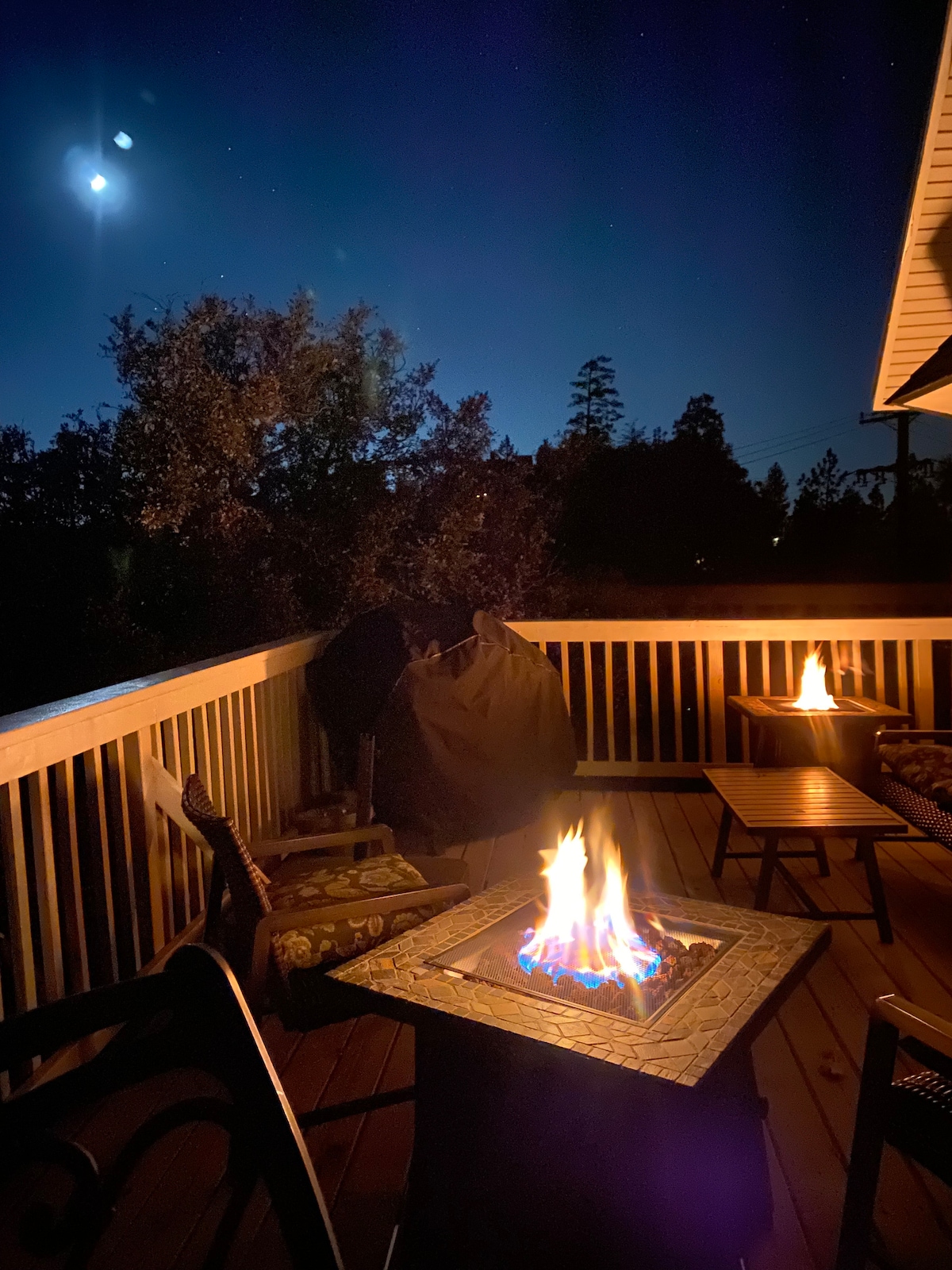 A cozy outdoor deck features a fire table with a bright flame, surrounded by seating. The night sky is visible overhead, showcasing stars and a hint of moonlight, while trees are softly illuminated in the background.