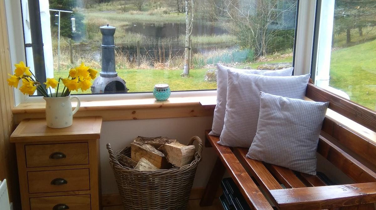 A sunlit corner seating area is shown with a wooden bench adorned with soft pillows. A rustic side table holds a small decorative candle and a vase of fresh daffodils. A woven basket filled with firewood sits nearby, with a view of the serene landscape visible through large windows.