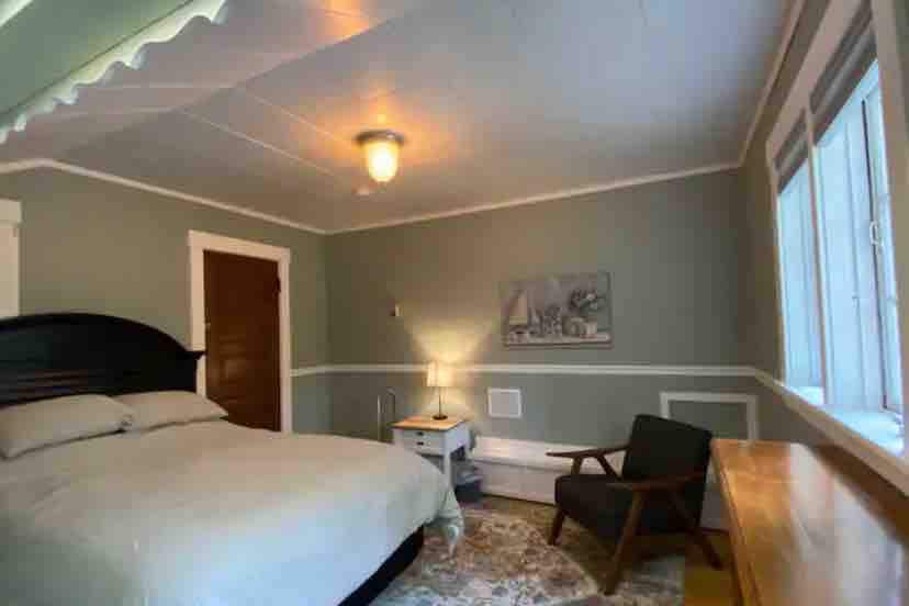 A spacious guest room features a queen size bed with light bedding. A nightstand and a chair accompany a wooden desk against the wall. Soft gray walls and natural light stream through multiple windows, creating a cozy and calming atmosphere.