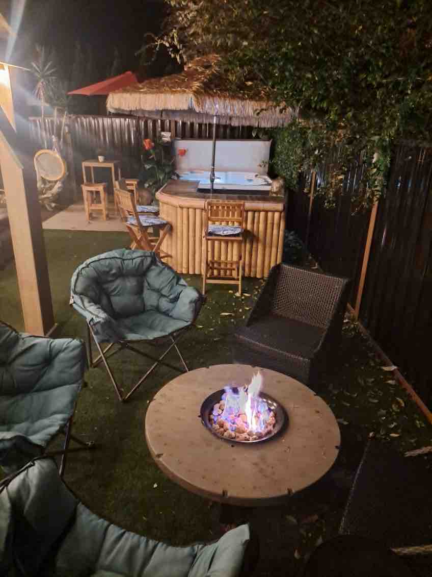 An outdoor space features a cozy seating area with several foldable chairs arranged around a circular fire pit. A hot tub is positioned in the background beneath an overhead structure, complemented by a small dining area with wooden stools.
