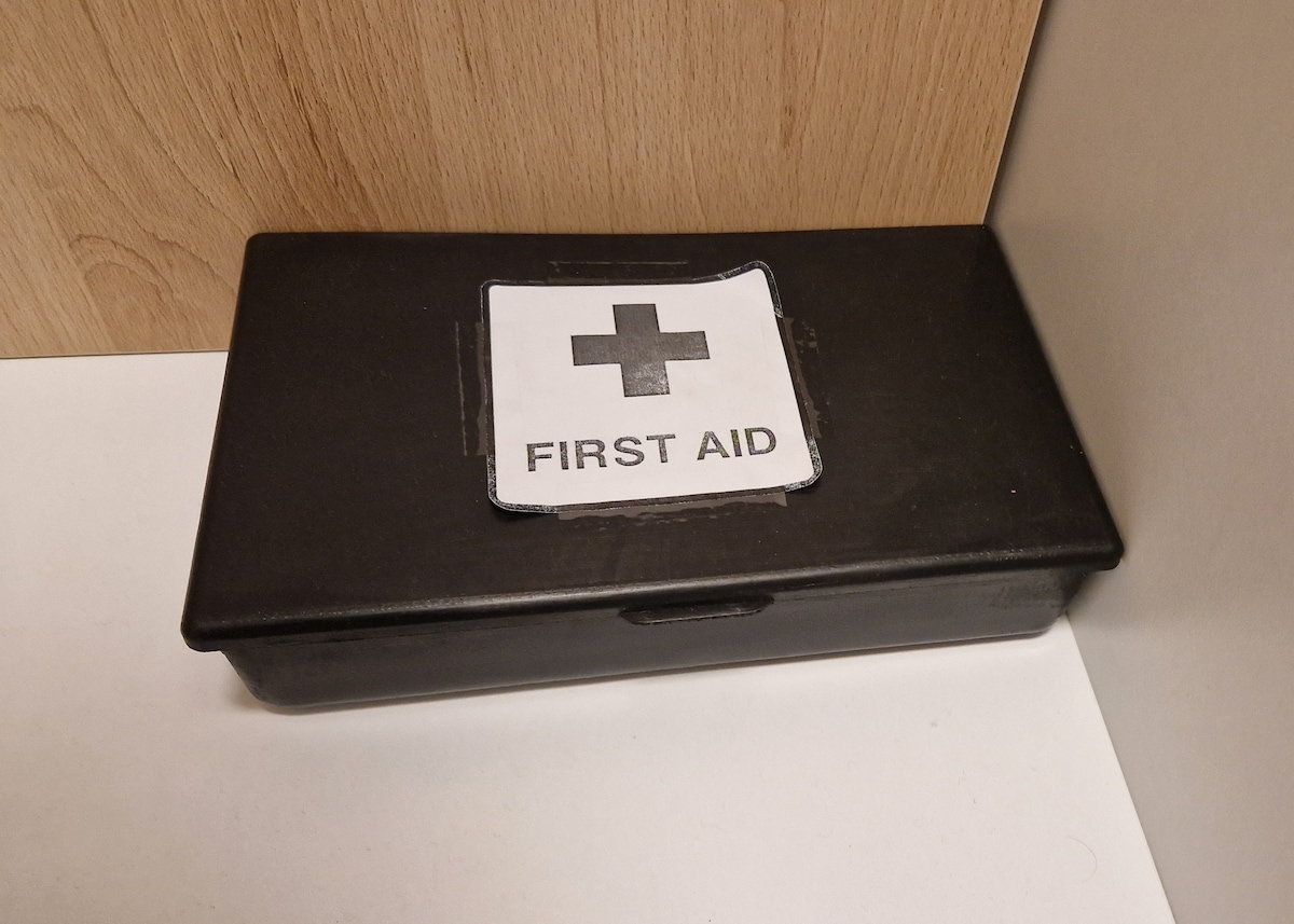A black first aid box rests on a white surface against a wooden background. The box features a clear label reading 'FIRST AID' with a red cross symbol, indicating its purpose for emergency medical supplies.