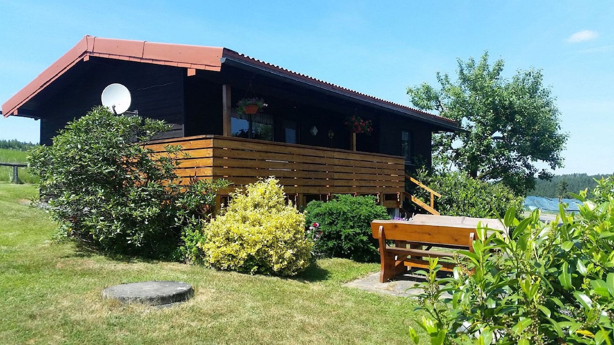 A charming wooden house with a red roof is surrounded by lush greenery and colorful flowers. A deck with wooden railings offers seating and a view of the adjacent garden, providing a peaceful outdoor space for relaxation.
