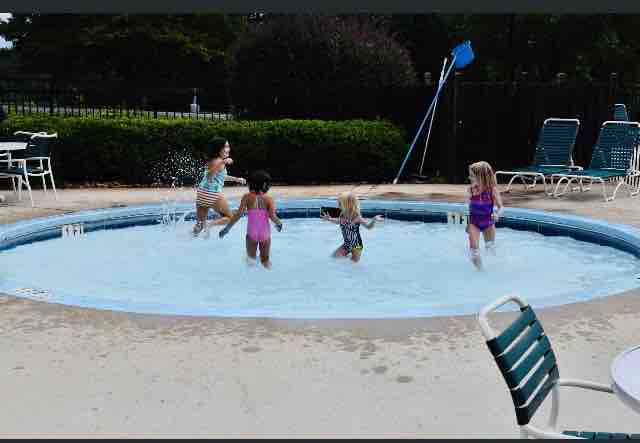 A circular pool area is shown with four children playing joyfully in the water. Surrounding the pool, green lounge chairs are arranged, and a blue poolside skimmer is seen in the background. Lush greenery borders the area, providing a serene atmosphere.