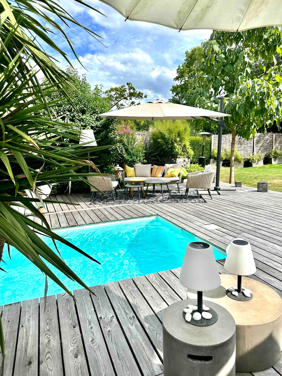 A private wooden terrace features a small swimming pool, surrounded by lush greenery. Cozy seating areas with cushions invite relaxation under a shaded pergola, while decorative lamps provide subtle lighting, enhancing the tranquil outdoor space.