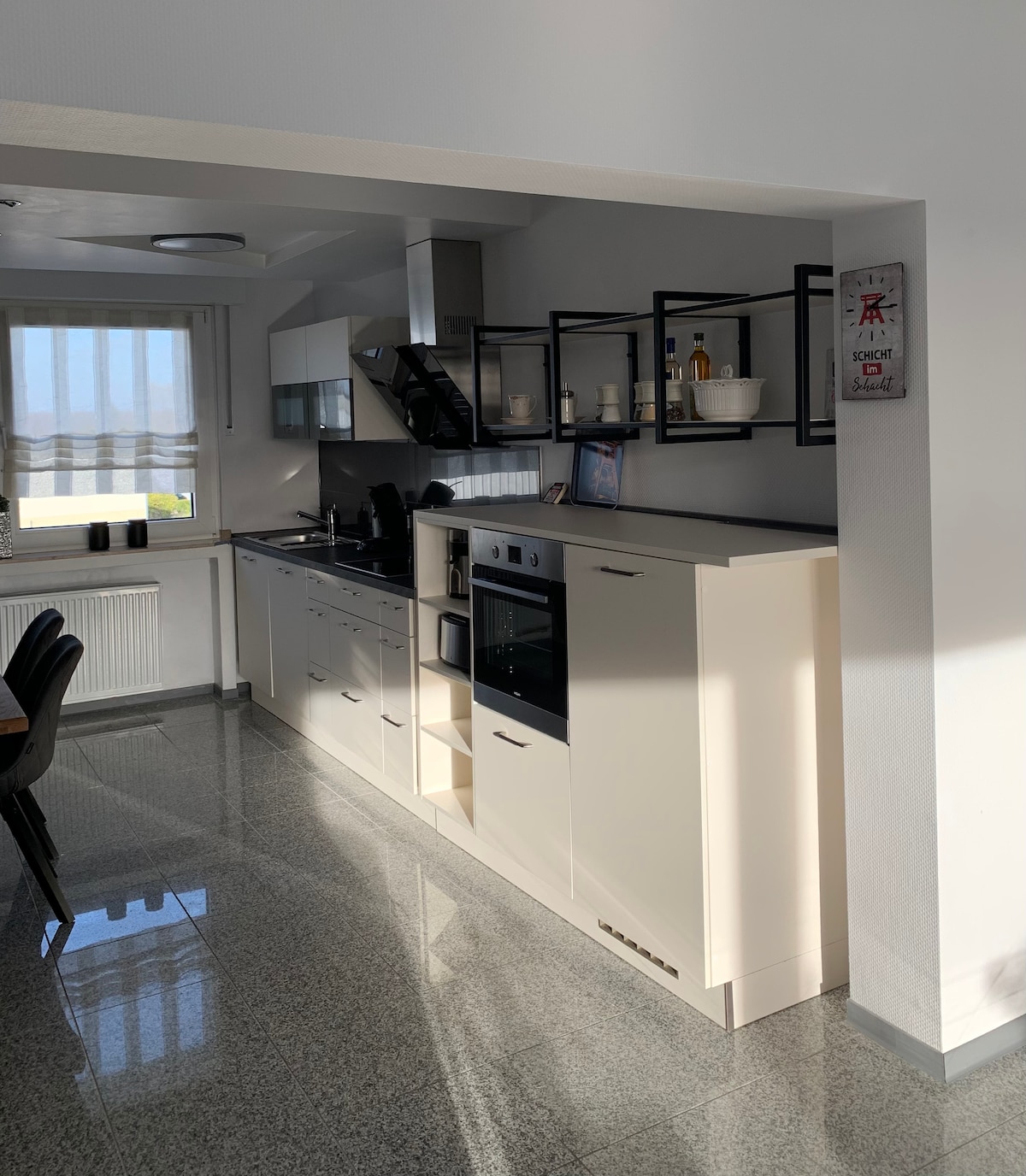 A modern kitchen is displayed, featuring sleek, white cabinetry and stainless steel appliances. A dark countertop aligns with the wall-mounted shelves, housing decorative items. The space is illuminated by natural light streaming through the nearby window, enhancing the clean and functional design.