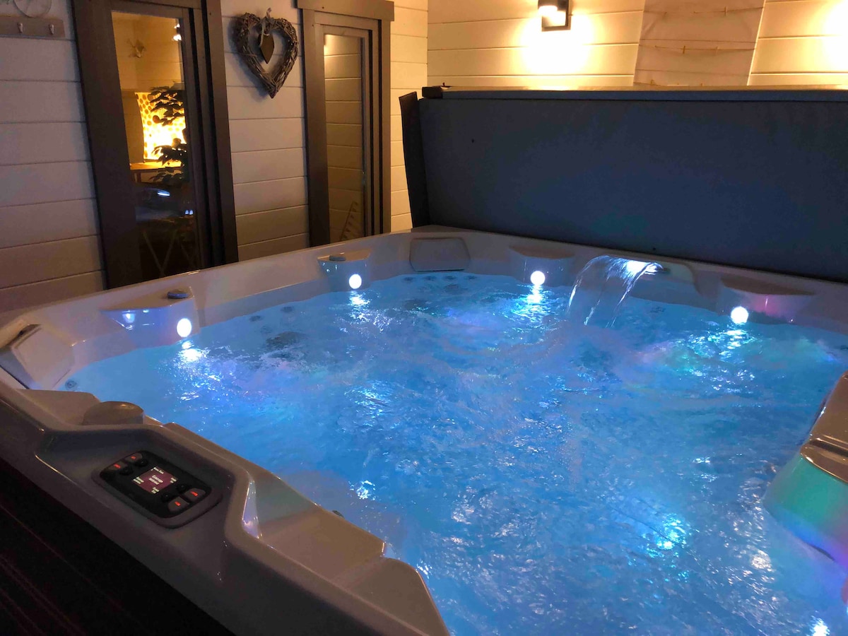 An inviting hot tub is featured, with cascading water creating a soothing ambiance. Soft blue lights illuminate the water, enhancing the overall relaxing environment. The area is surrounded by wooden walls and large glass doors, providing a view into the living space.