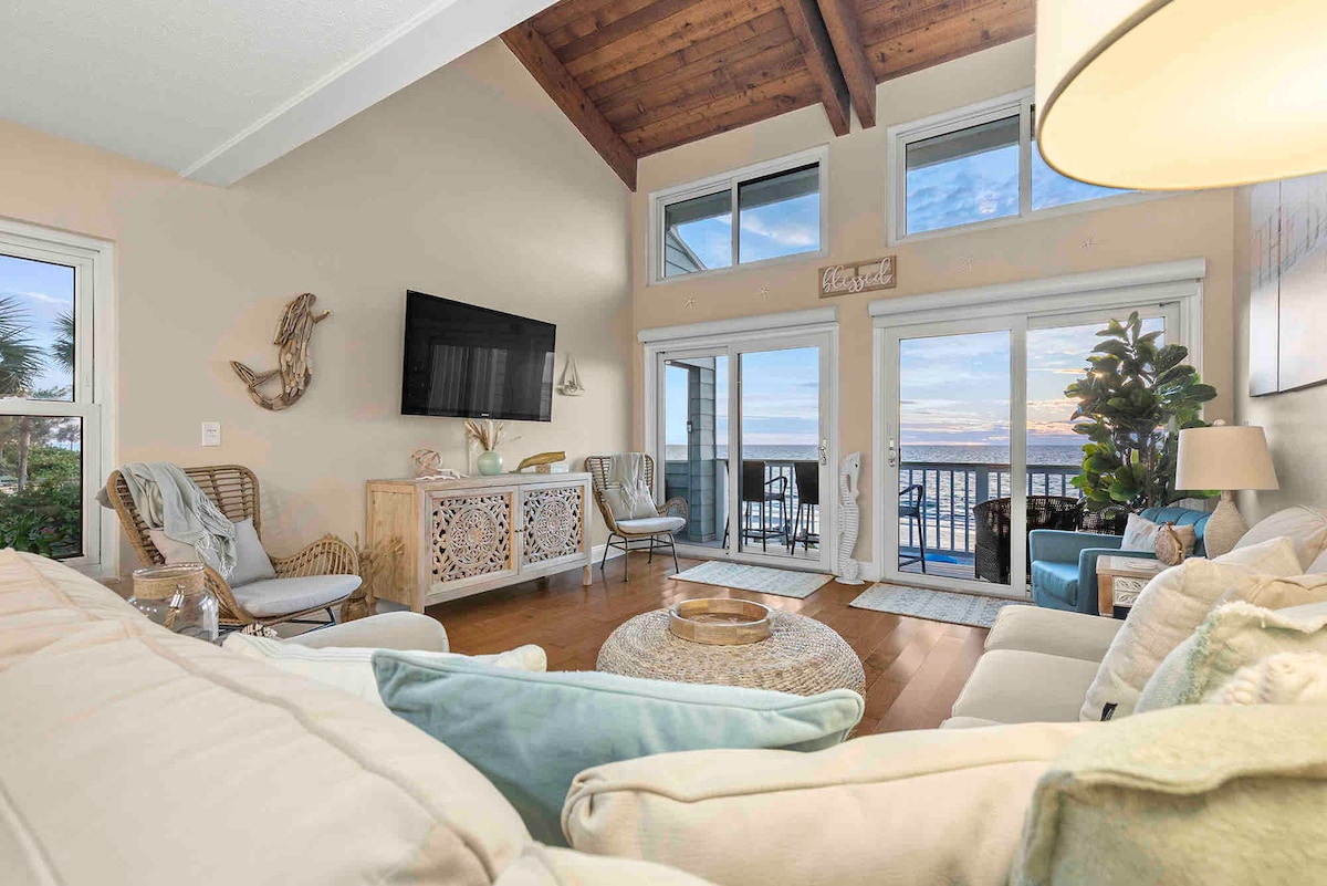 The living room showcases a spacious layout with large windows offering ocean views. Natural light floods the room, highlighting the wooden beams of the ceiling. Comfortable seating arrangements are evident, complemented by soft decor elements, creating a serene atmosphere consistent with coastal living.
