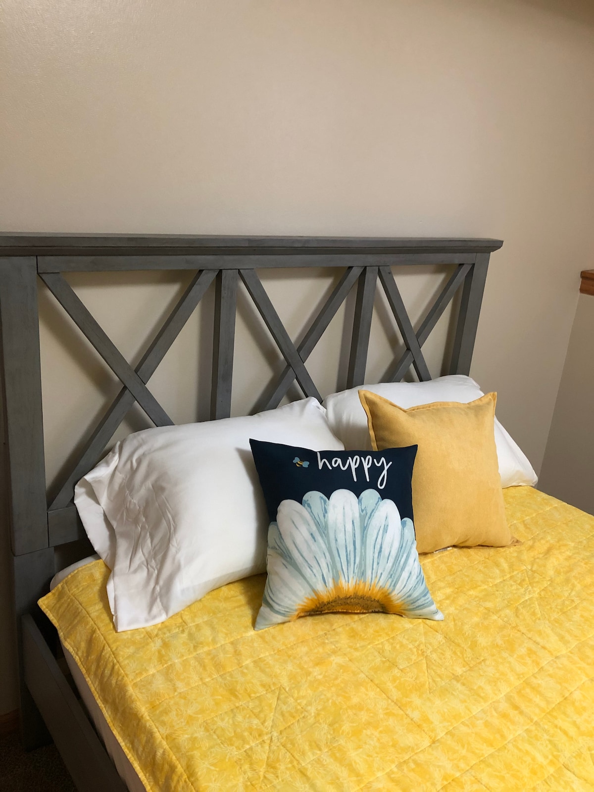 A modern bed frame with a geometric headboard is paired with soft, white pillows and a coordinating yellow quilt. Two accent pillows, one featuring a daisy design and the word 'happy,' add a touch of color to the inviting sleeping area.