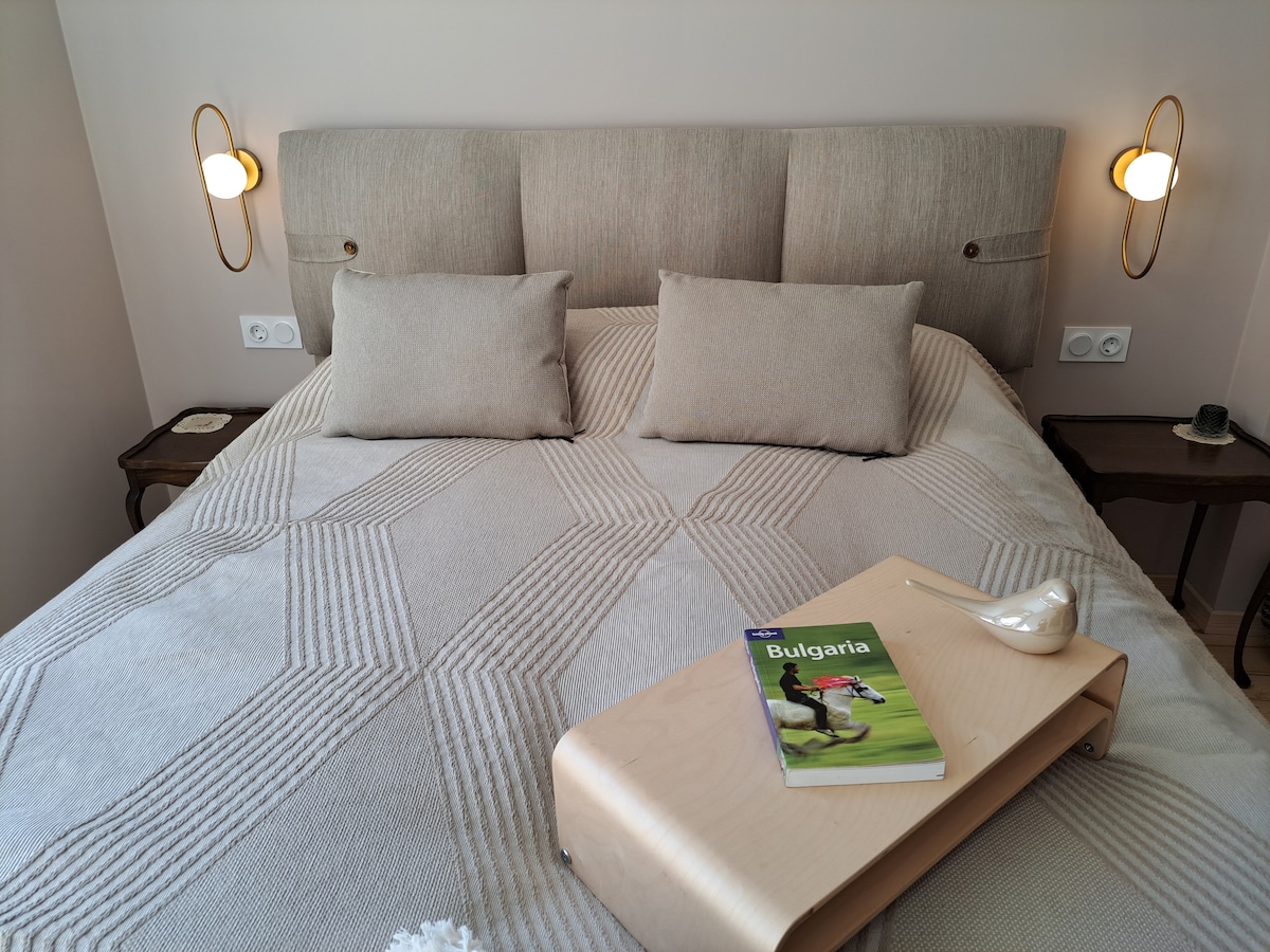 A spacious bed is dressed in neutral-toned bedding and accented by two soft pillows. A minimalist side table holds a brochure titled 'Bulgaria' and a decorative object. Warm lighting fixtures are mounted on the wall beside the bed.