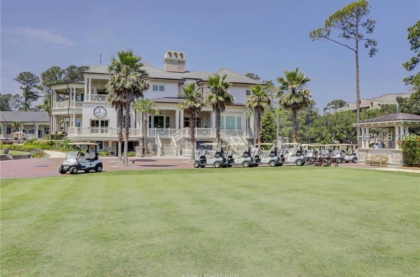 A grand clubhouse sits surrounded by lush landscaping, featuring palm trees and manicured lawns. Golf carts are lined up neatly in front of the building, showcasing easy access to the golf course. A large clock adorns the exterior, signaling a welcoming atmosphere.